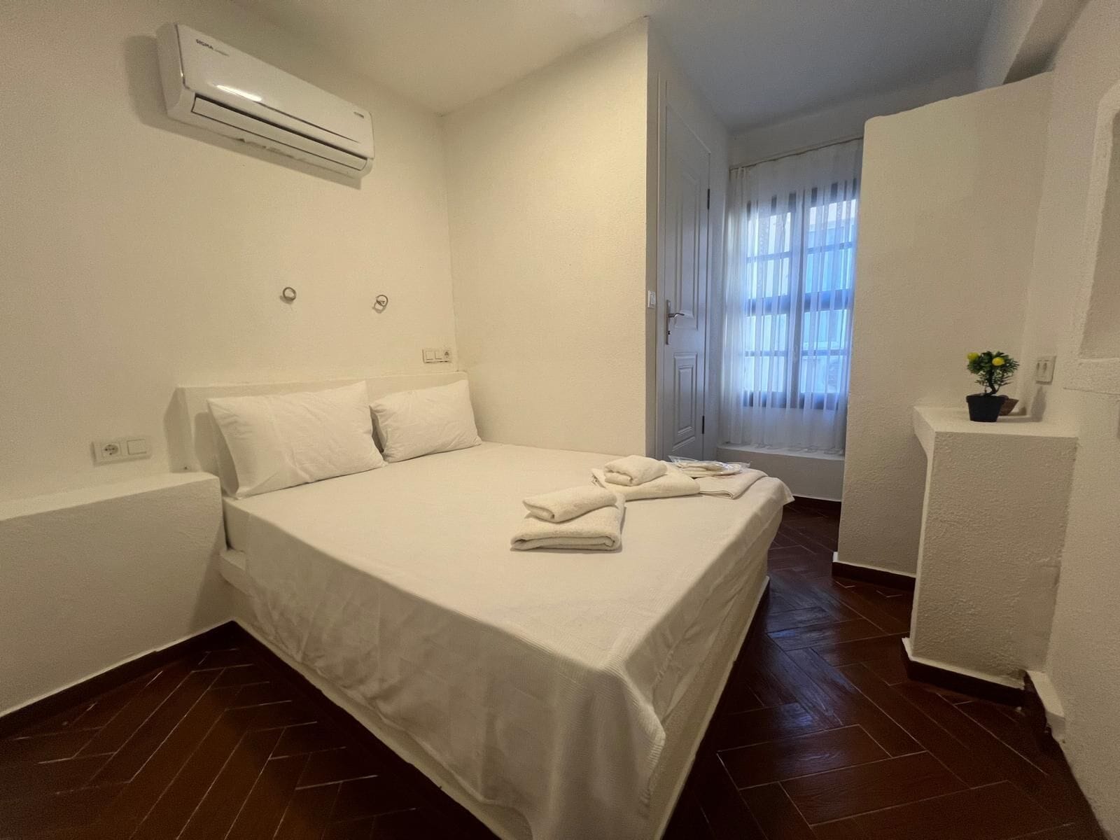 A hotel room with a bed, white walls, an air conditioner, a window with curtains, and a small plant on a shelf.