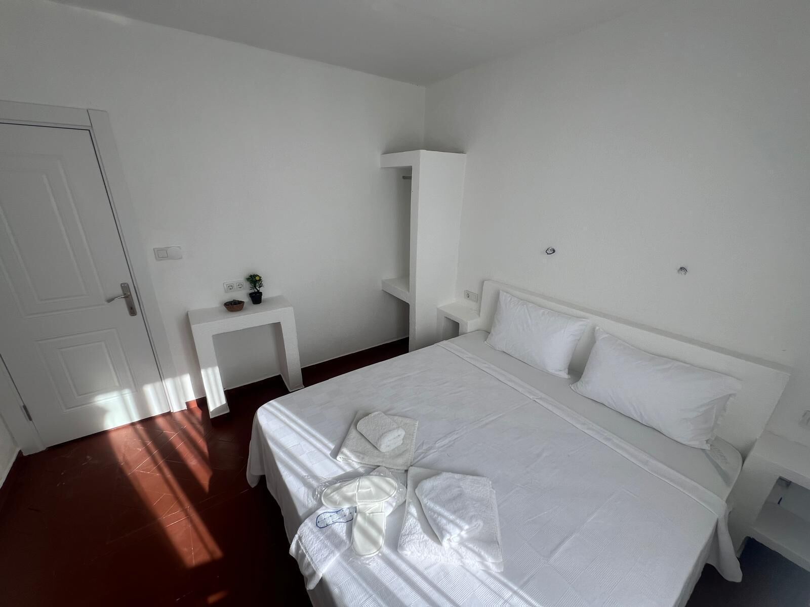 A white room with a bed, a small table with a plant, and a door.