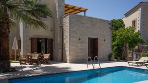 A swimming pool with a stone wall and wooden doors, surrounded by outdoor furniture and a palm tree.