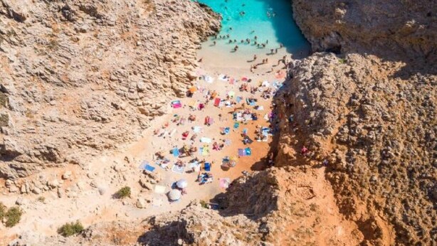 Eine felsige Küstenlinie mit einem Strandabschnitt, der zahlreiche Sonnenschirme und Personen zeigt.