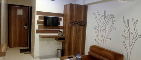 A modern hotel room with a wooden wardrobe, a flat-screen TV, a sofa, and a wall with tree line art.