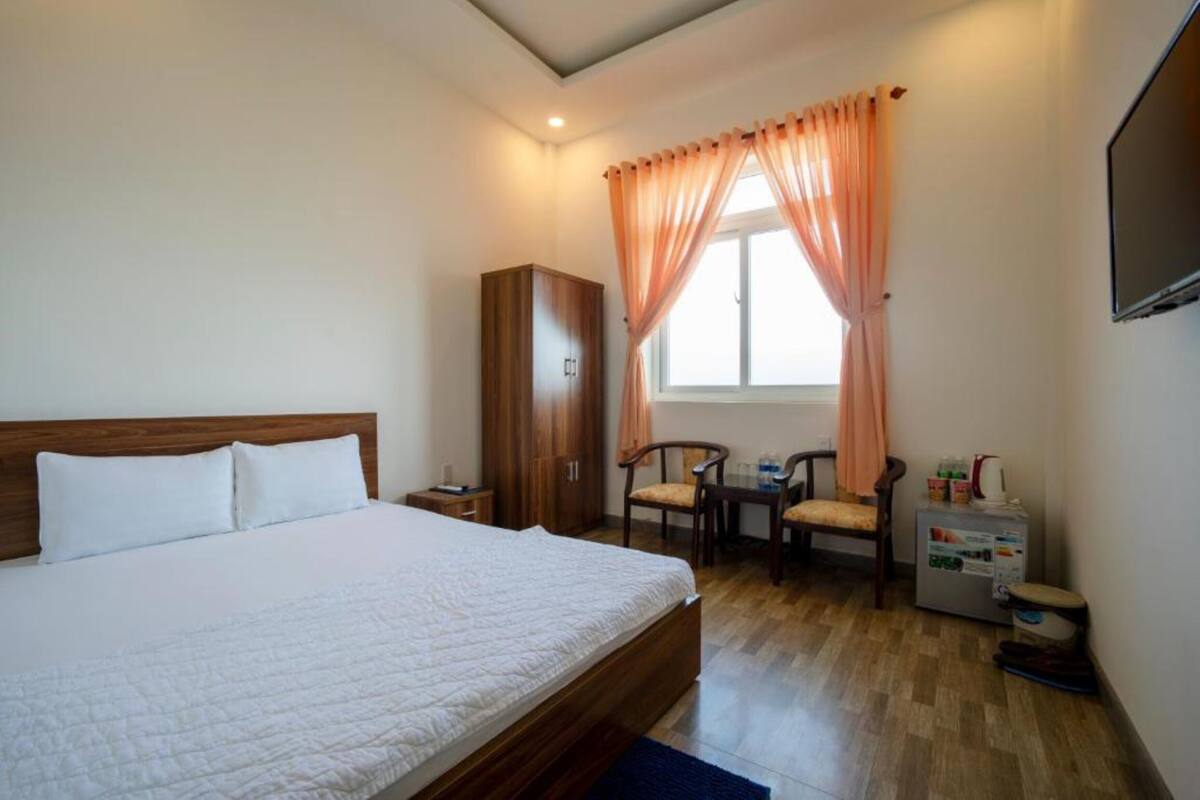 A hotel room with a large bed, wooden wardrobe, a small table with chairs, a window with curtains, and a television mounted on the wall.