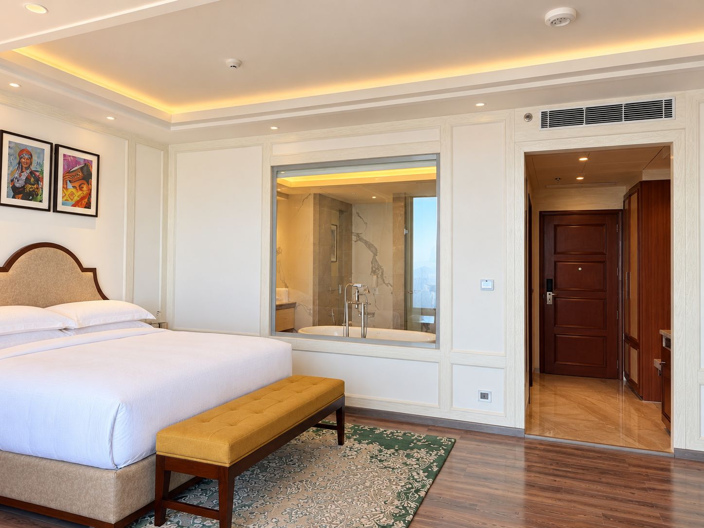 A hotel room with a large bed, a bench, a bathroom with a glass shower enclosure, and a door leading to another room.