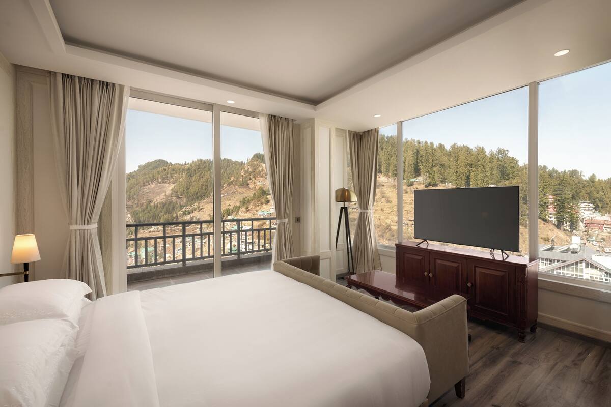 Une chambre d’hôtel dotée d’un grand lit, d’une télévision à écran plat, d’un bureau et d’un balcon offrant une vue sur les collines.
