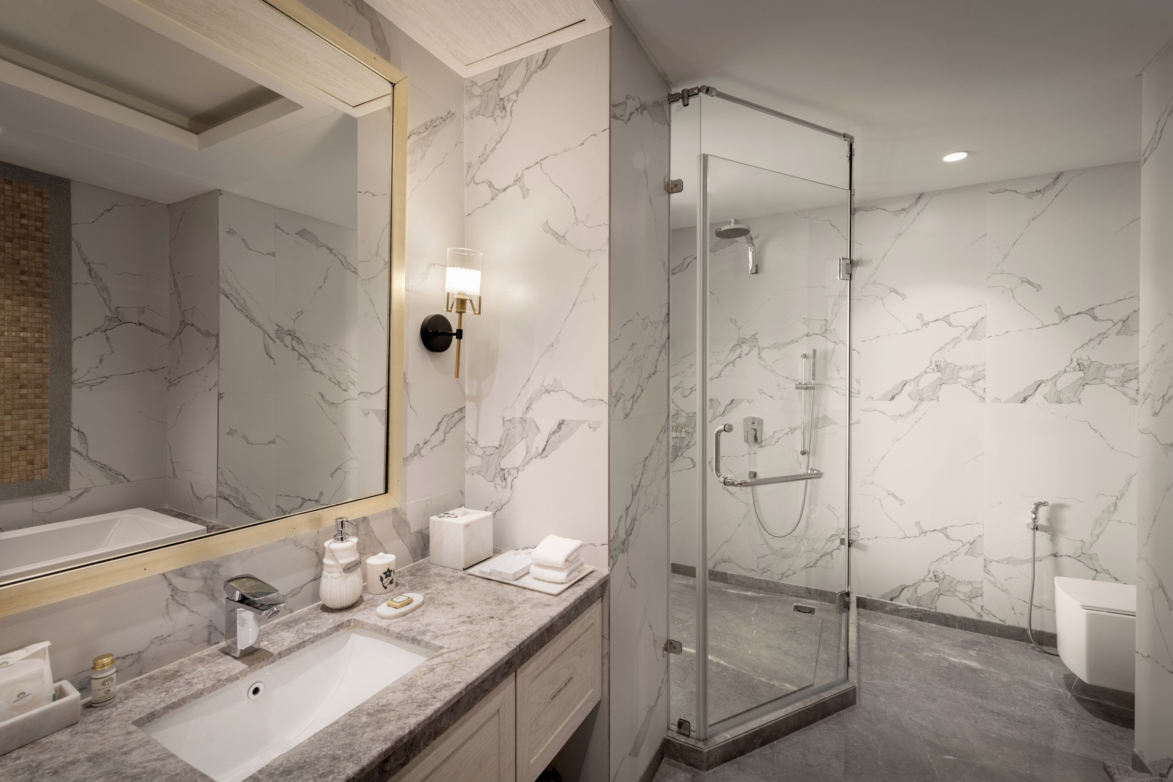 A modern bathroom with a marble wall, a large mirror, a sink, a shower, and a toilet.