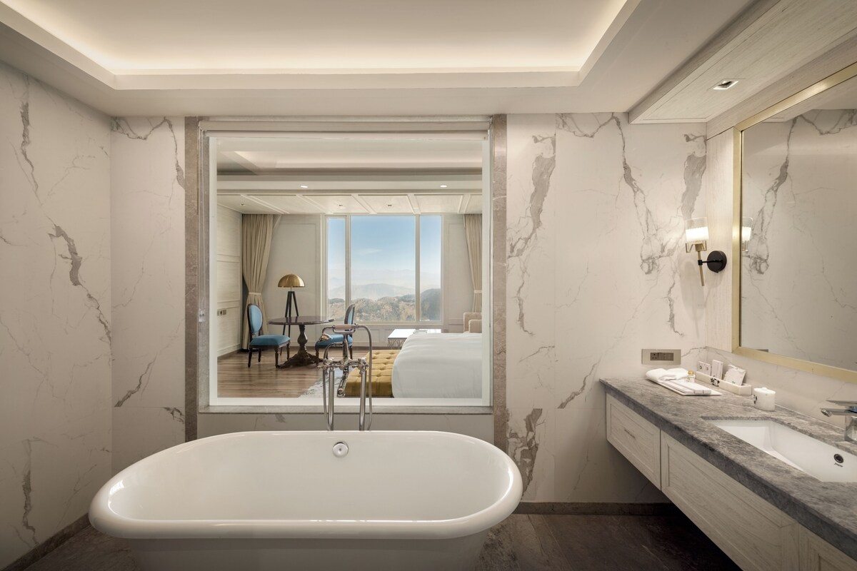 Une salle de bain moderne avec une baignoire sur pieds, des murs en marbre, un grand miroir et une vue sur une chambre à coucher avec un lit et un fauteuil.