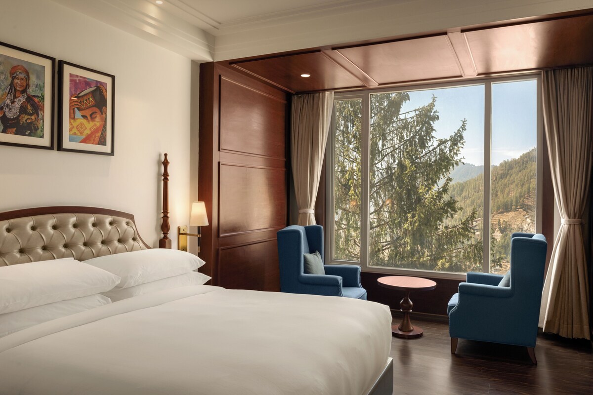 Une chambre d’hôtel avec un grand lit, deux fauteuils, une petite table et une fenêtre donnant sur des arbres et des collines.