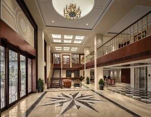 Eine geräumige Lobby mit einem prächtigen Kronleuchter, einem Schachbrettmuster auf dem Boden und einer Treppe, die zu einer oberen Ebene führt.