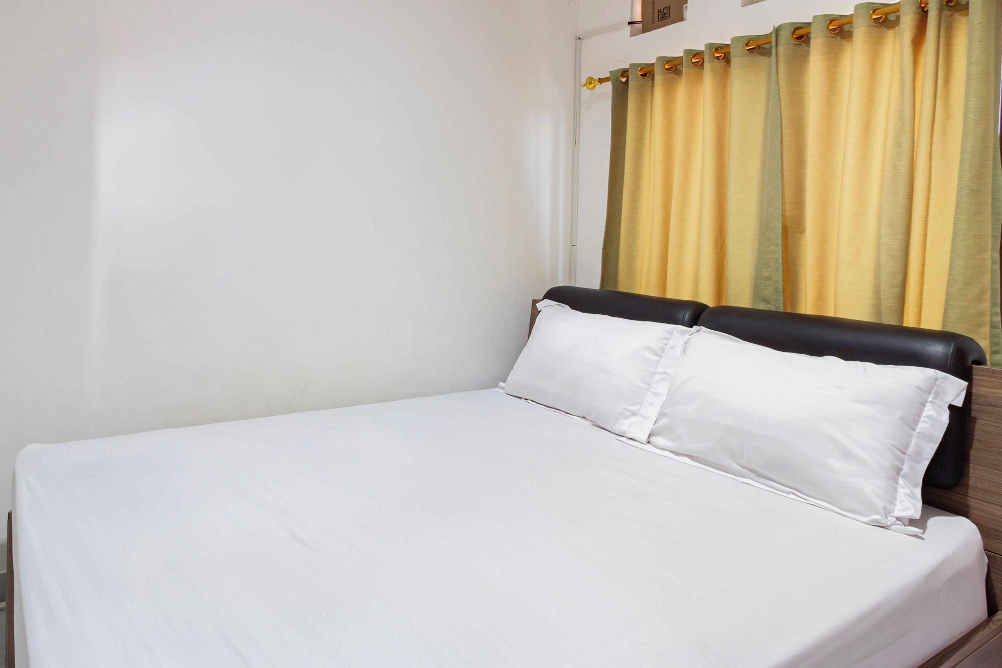 A single bed with white bedding in a room with a plain wall and a curtain with a yellow and green pattern.