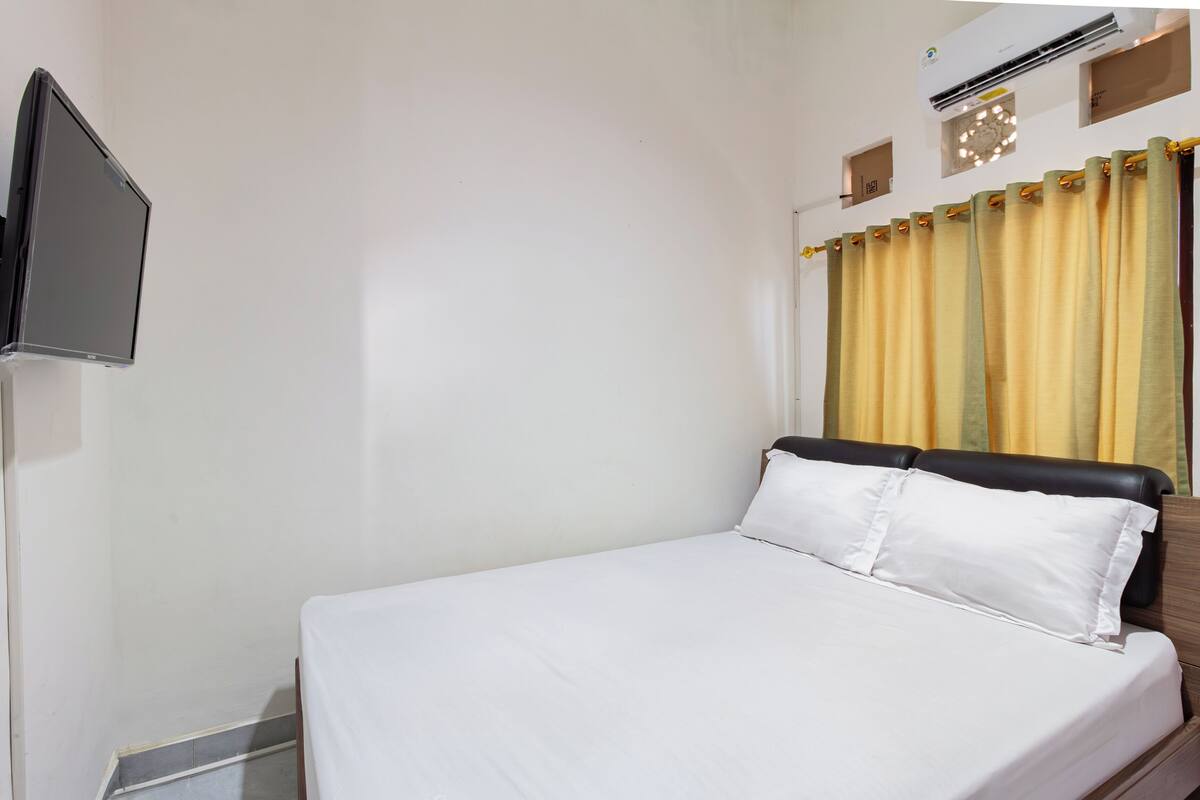 A neatly made bed with white linens, a flat-screen TV mounted on the wall, and a window with yellow curtains.