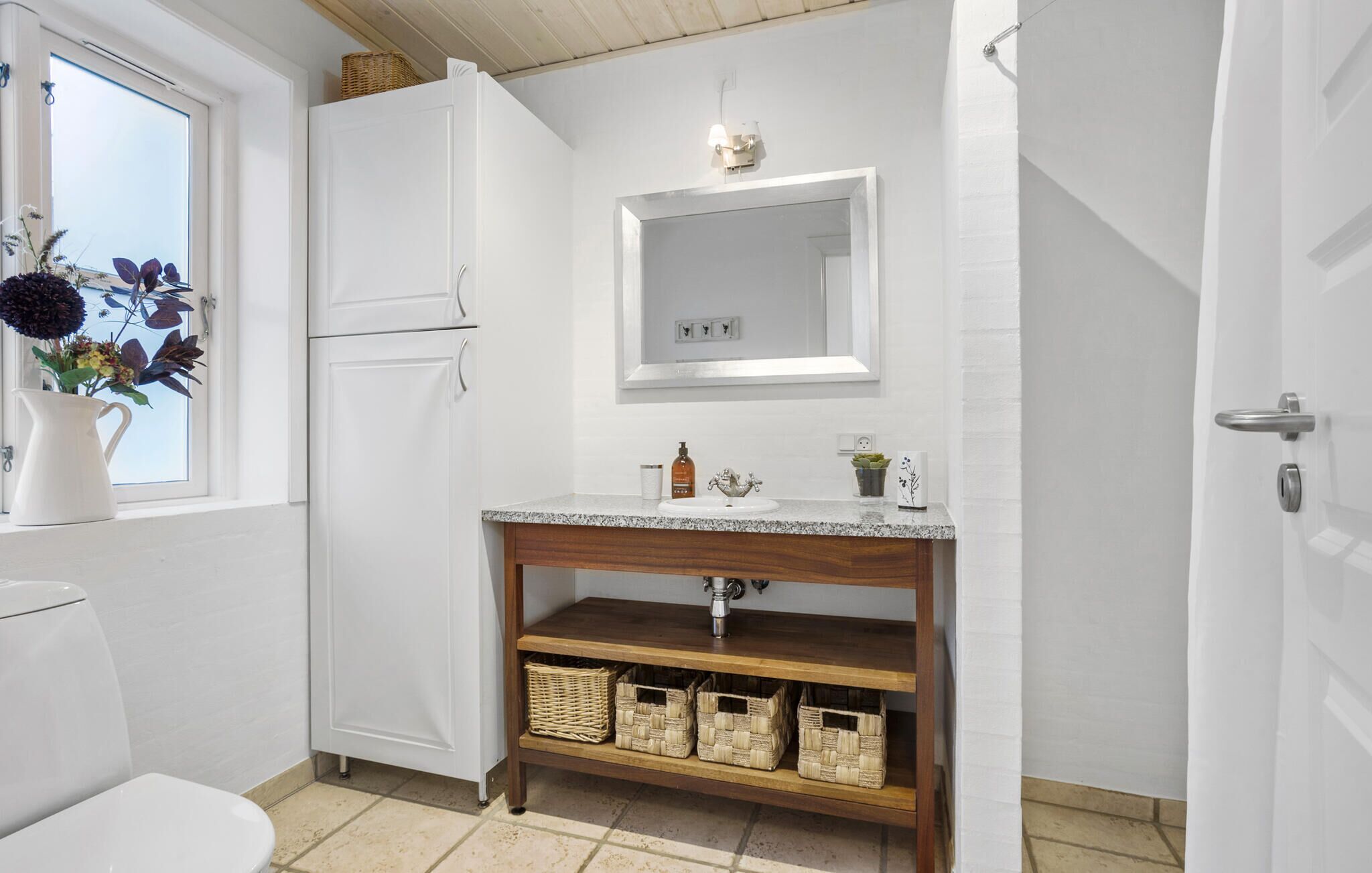 A bathroom with a white toilet, a sink with a wooden vanity, a mirror, and wicker baskets on the shelf.
