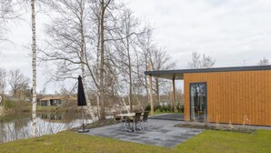 A modern building with a wooden exterior, a dining area with chairs and a table, and a pond in the background. - Lakefront Holiday Home (Hoge Hexel)