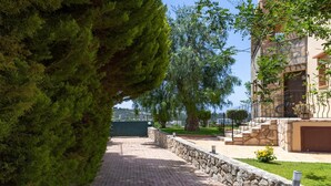 A paved pathway leading to a stone wall, with a large tree on the left and a building with a stone facade and a balcony on the right. - Villa Pasiphae 1 in Stalos (Chania)