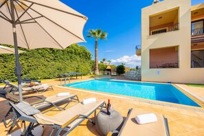 A swimming pool with lounge chairs and umbrellas, surrounded by a well-maintained hedge and a building with balconies. - Villa Pasiphae 2 in Stalos (Chania)