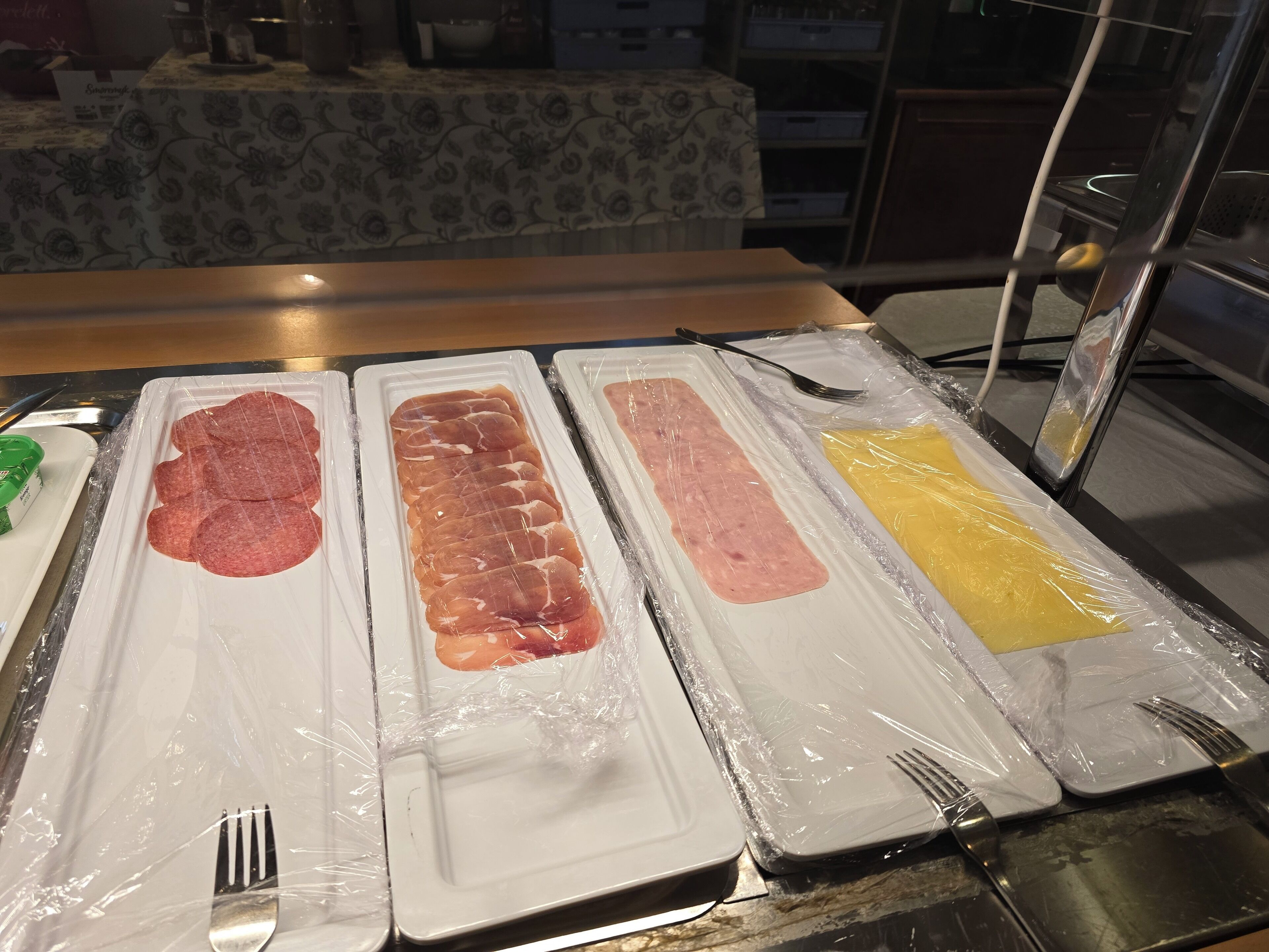 A buffet table with various food items wrapped in plastic, including sliced meats and a yellow dish, possibly a dessert.