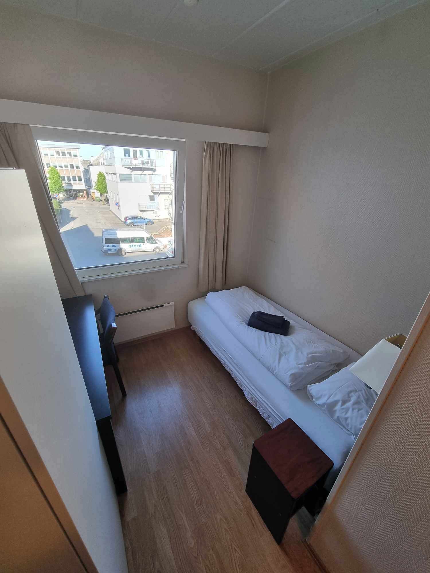 Ett litet rum med en enkel säng, ett fönster, ett skrivbord och en stol.