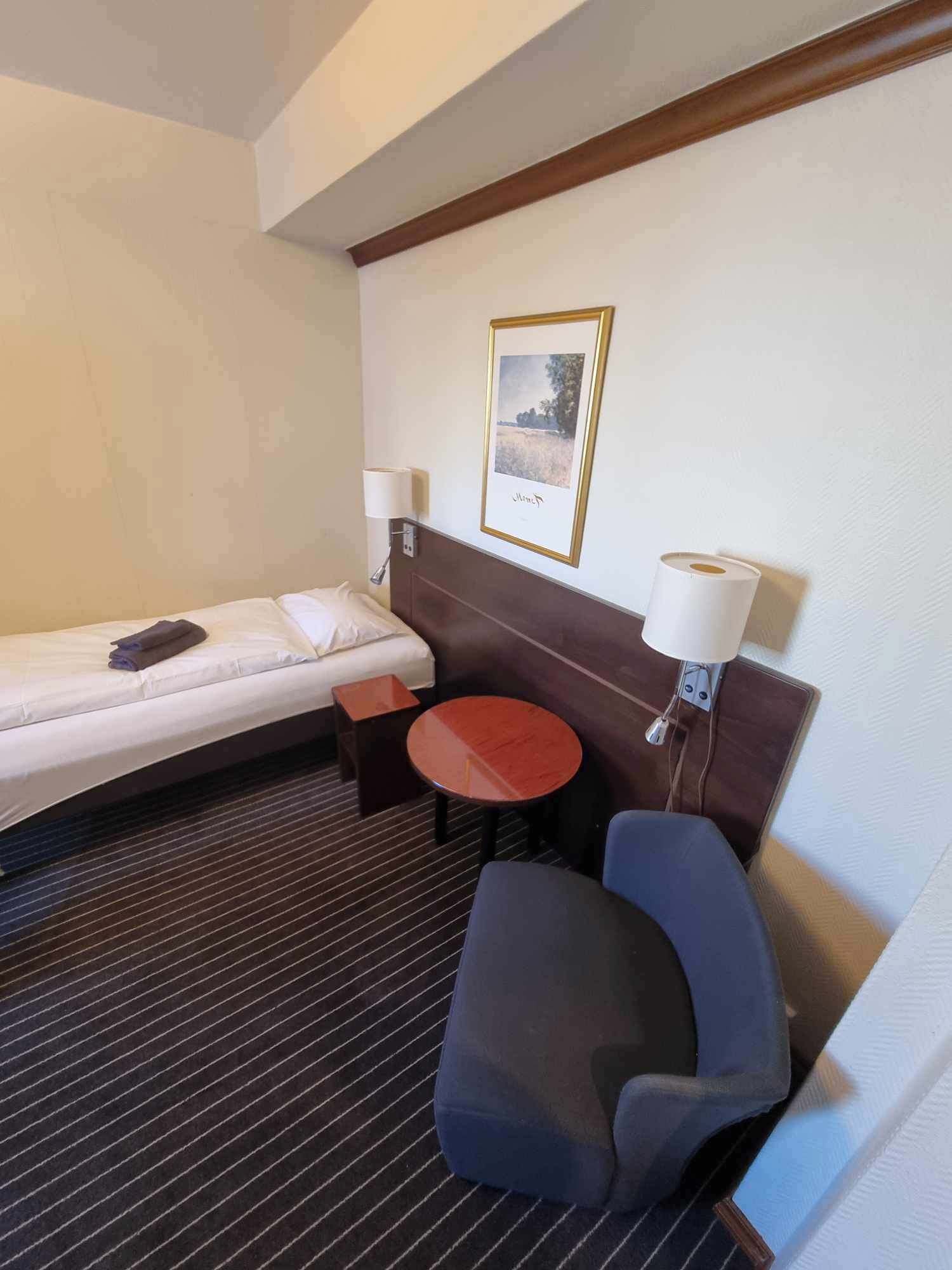 Una habitación de hotel con una cama, una mesita, una silla y una lámpara fijada a la pared.