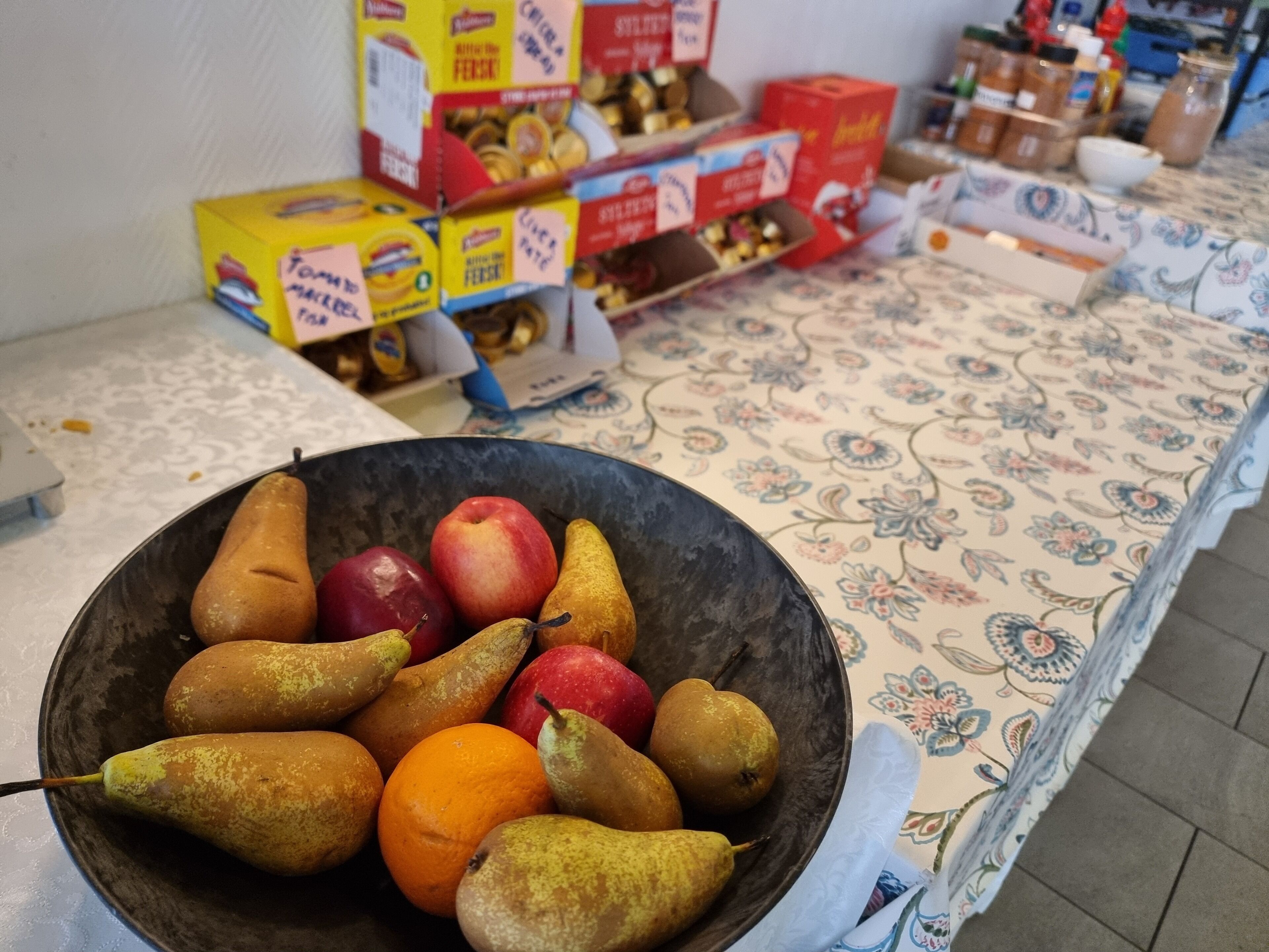 A table with a bowl of fruit and boxes of cereal in the background.