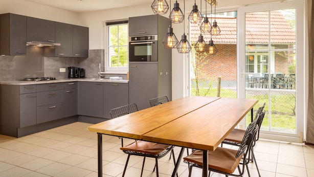 Een moderne keuken met grijze kastjes, een houten eettafel en een groot raam met uitzicht op een bakstenen gebouw.