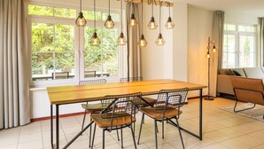 Een eetgedeelte met een houten tafel, metalen stoelen, een hanglamp en uitzicht op groen door grote ramen.