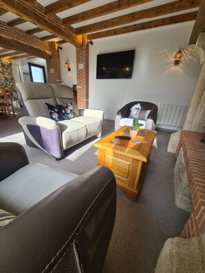 En stue med sofa, lænestol og sofabord, et fjernsyn monteret på væggen og en murstenspejs.