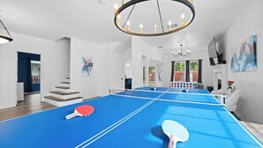 A spacious room with a ping pong table, two ping pong paddles, a chandelier, and a staircase leading to another area. - Modern 5 BR Sleeps 16 with Pool, Spa, Air Hockey, BBQ & Games Near Downtown! (San Antonio)