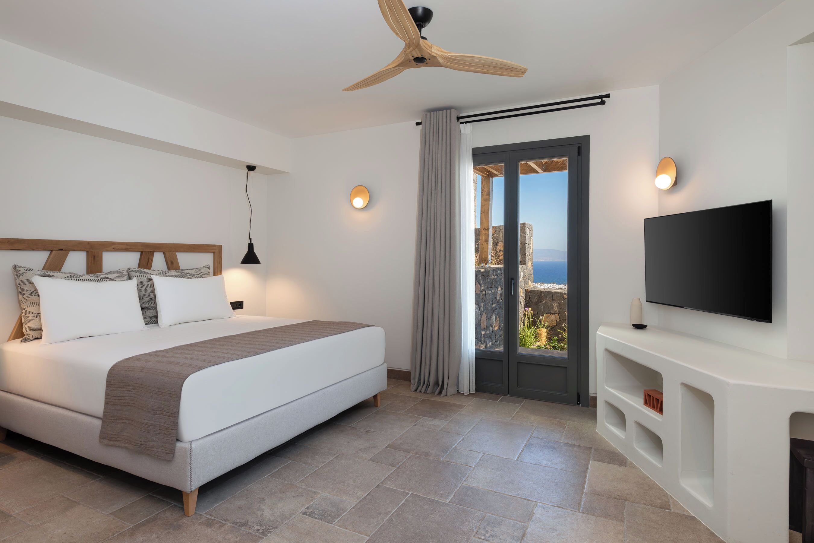 A modern hotel room with a large bed, a ceiling fan, a TV mounted on the wall, and a balcony with a view.