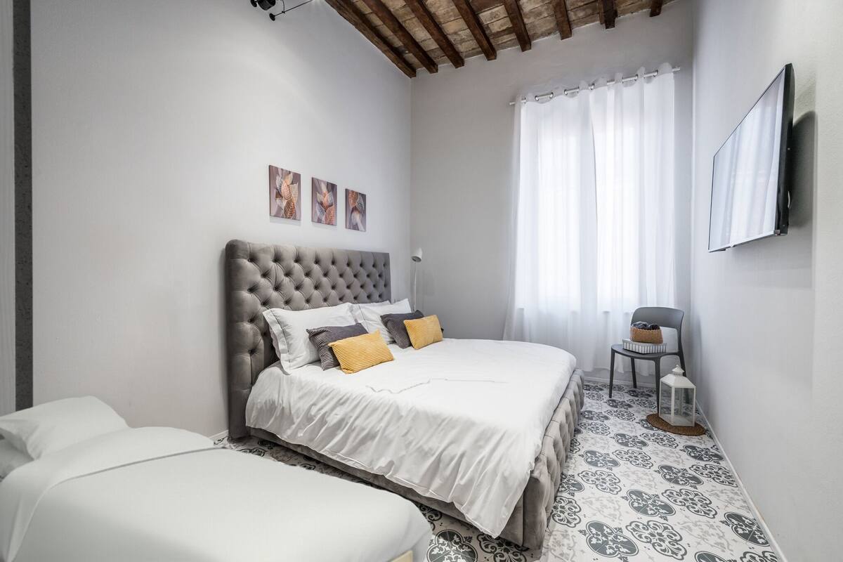 A bedroom with a tufted headboard, a bed with white bedding, a bedside table, a chair, and a TV mounted on the wall.