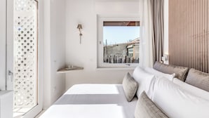 A modern bedroom with a large bed, a window with a view of buildings, a small bedside table, and a wall-mounted lamp. - Diff'rent Souls Rooms (Sorrento)