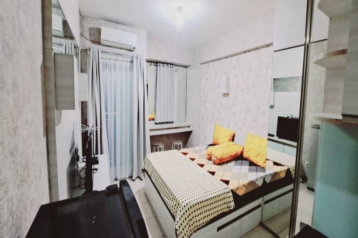 Quarto pequeno e acolhedor, com cama, escrivaninha e cadeira. Há uma janela com cortinas, um radiador e uma mesinha.