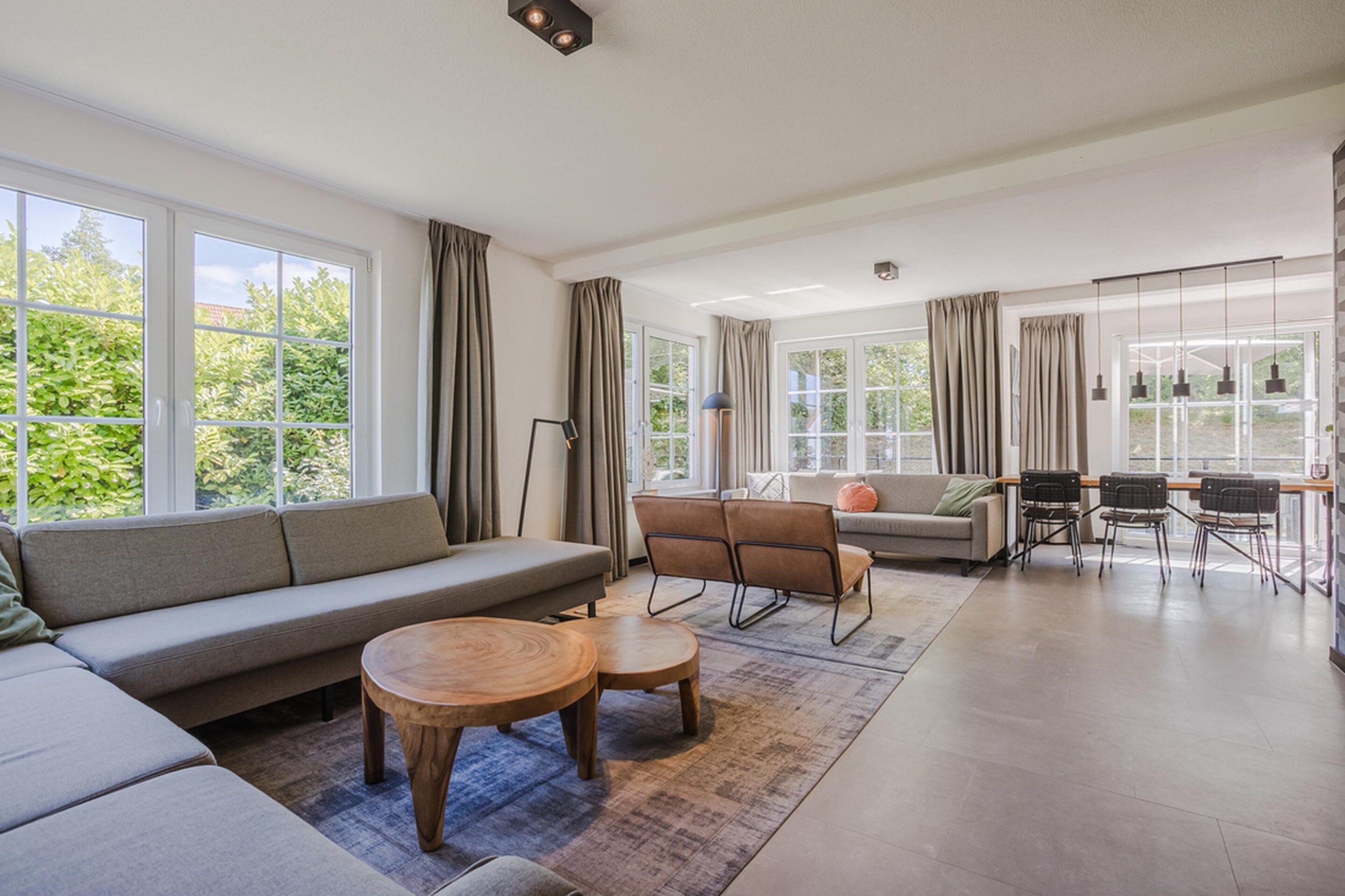 Amplia sala de estar con sofá, sillones, una mesa de centro y un comedor con sillas y mesa. Amplias ventanas ofrecen luz natural y vistas a zonas verdes.