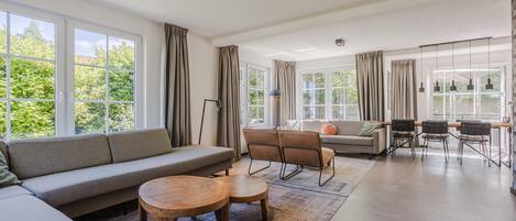 A spacious living area with a sofa, chairs, a coffee table, and a dining area with chairs and a table. Large windows provide natural light and views of greenery outside.