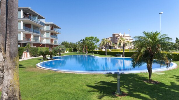 A swimming pool surrounded by well-maintained grass and palm trees, with a multi-story building in the background. - Happy in Oliva With 1 Bedrooms and 1 Bathrooms (Oliva)