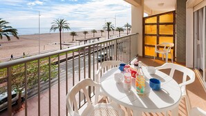 A balcony with white plastic chairs and a table, overlooking a beach with palm trees and a road with cars. - Infante in Grau i Platja (Gandia)