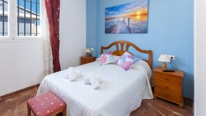 A neatly made bed with white linens and multiple pillows, a wooden nightstand with lamps, a bench with a floral cushion, and a sunset painting on the wall. - Golden in Gandia With 3 Bedrooms and 3 Bathrooms (Gandia)