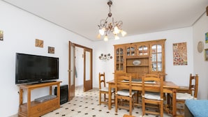 A living room with a TV on a wooden stand, a dining table with chairs, a chandelier, and patterned wall decorations. - Fuentes Del Colorao in Barrio Nuevo (Conil de la Frontera)
