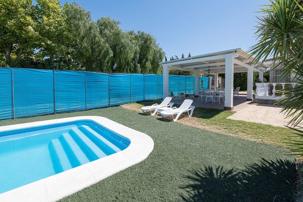 A swimming pool with a white deck, surrounded by a blue fence. There are white lounge chairs and a covered patio area with tables and chairs.