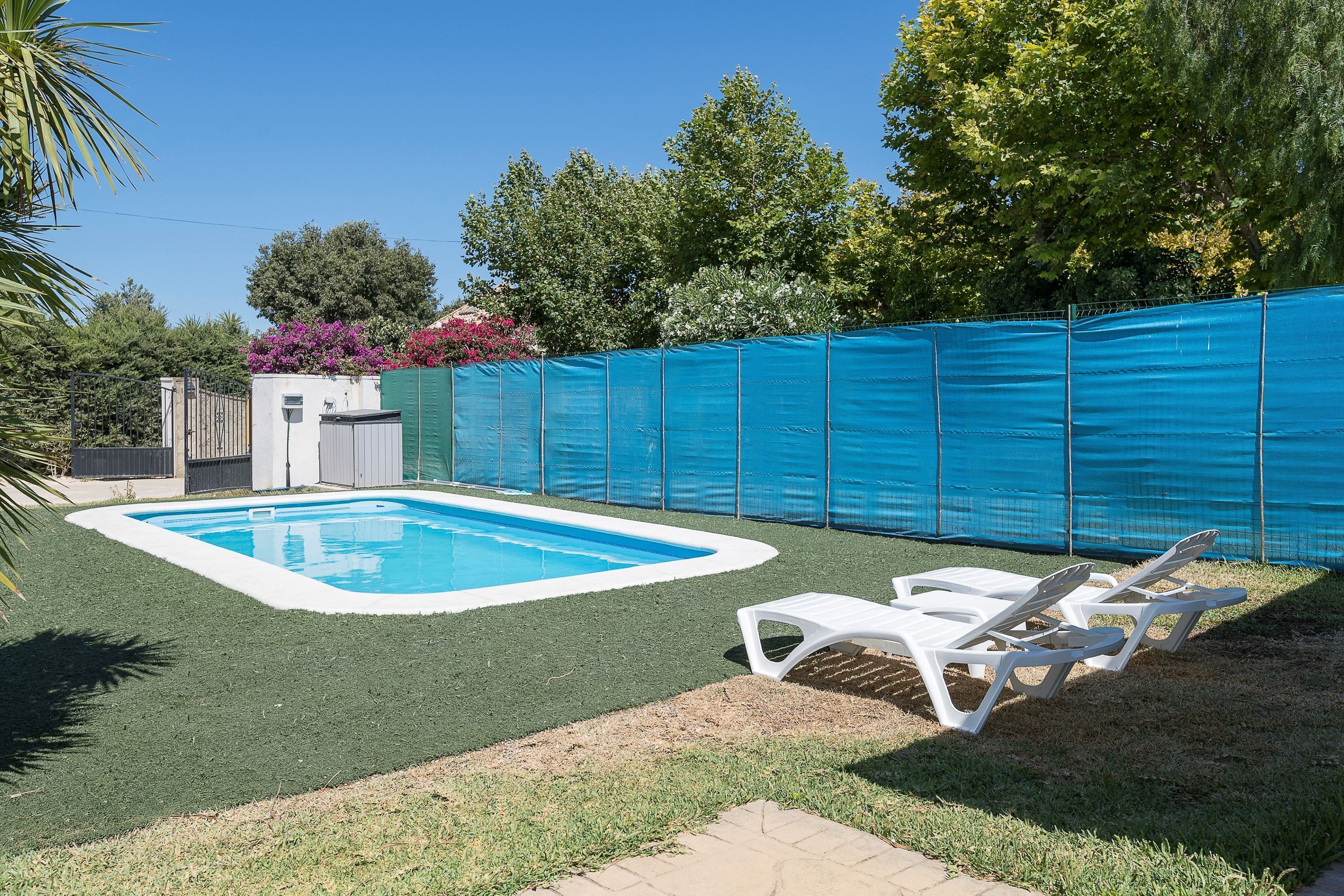 Una piscina con zona de césped artificial verde, una cerca y dos sillas reclinables blancas.