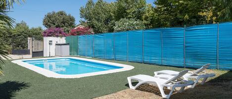 Una piscina con zona de césped artificial verde, una cerca y dos sillas reclinables blancas.