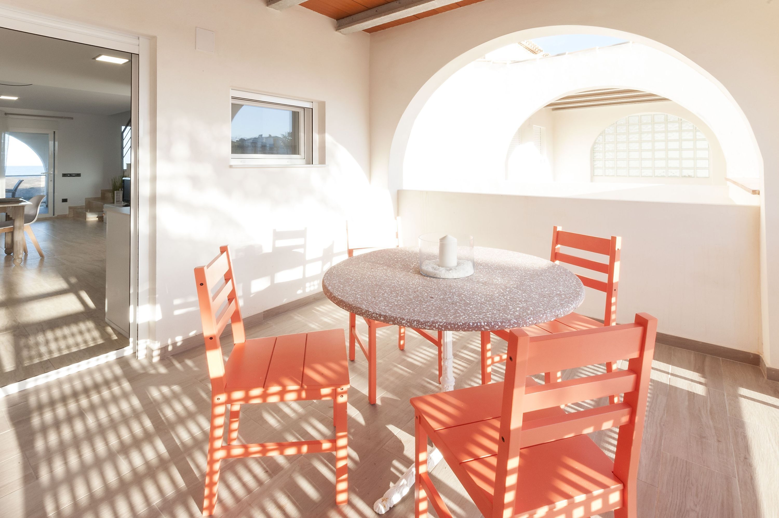 Una habitación con una mesa redonda y cuatro sillas naranjas, una ventana con vistas y una puerta que da a otra habitación.