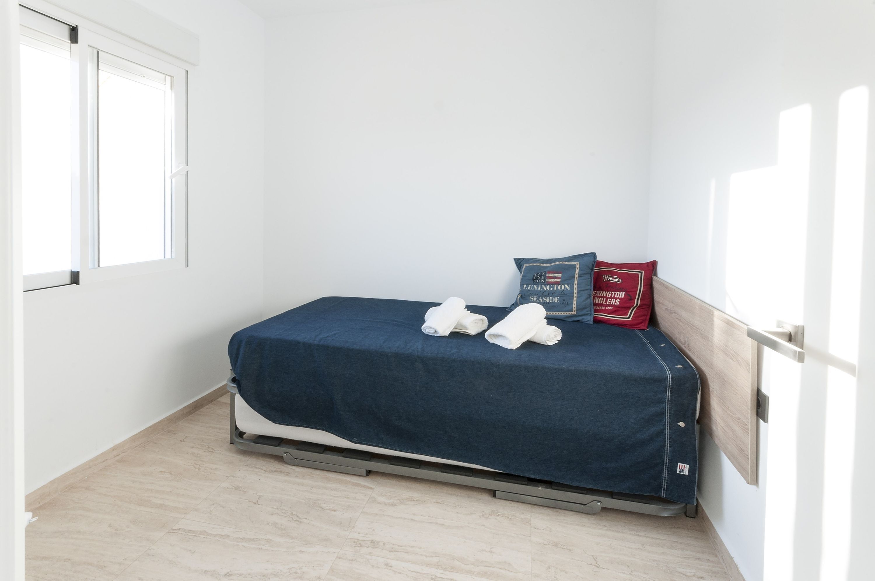 A small, neatly made bed with a blue cover, two pillows, and a red pillow with a logo.