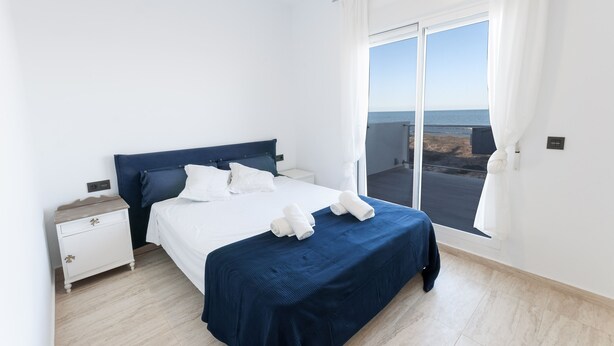 Un dormitorio con una cama grande, mesitas de noche blancas y vista al mar a través de un ventanal amplio.