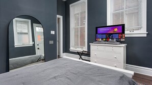 Une chambre à coucher avec un lit, un téléviseur posé sur une commode et une fenêtre avec des stores.