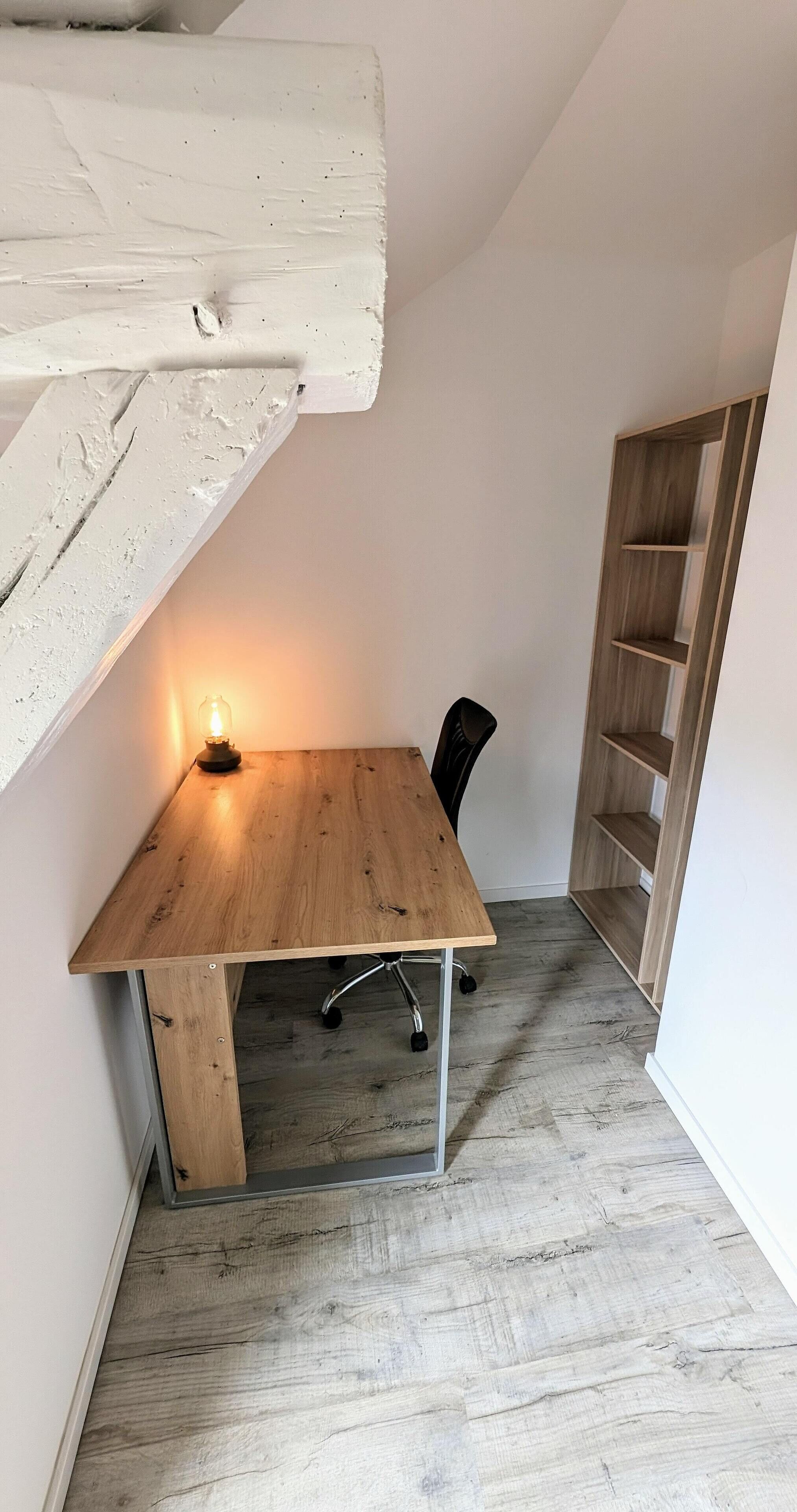 Habitació d'angle amb escriptori de fusta, una cadira, una làmpada de taula petita i un moble prestatge de fusta encastrat.