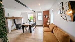 A modern living room with a wooden floor, a brown sofa, a dining table with chairs, a fireplace, and a large window. - Maison Lac et Jardin (Mesnil-Saint-Père)