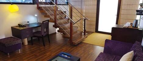 A modern living room with a wooden staircase, a desk, a purple sofa, and a coffee table with fruits.