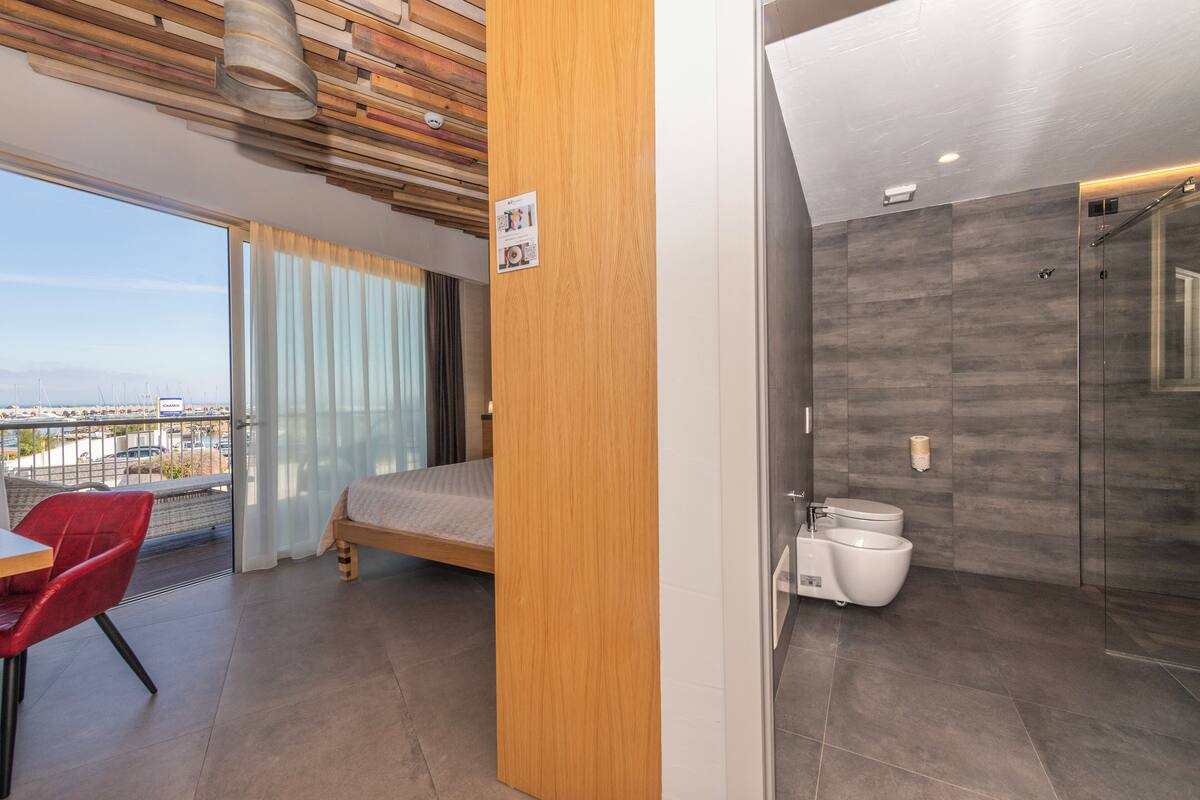 A modern hotel room with a balcony, a bed, a shower area, and a dining area with a red chair.