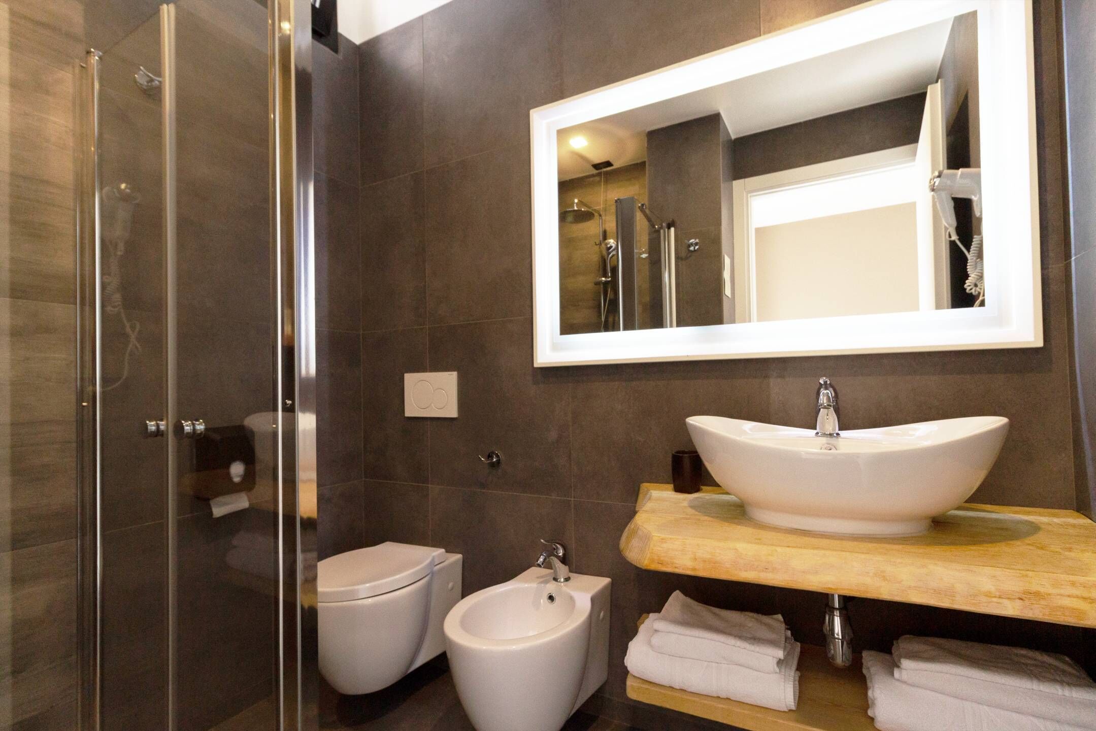 A modern bathroom with a glass-enclosed shower, a white toilet, a bidet, and a large mirror above a wooden vanity with a sink.