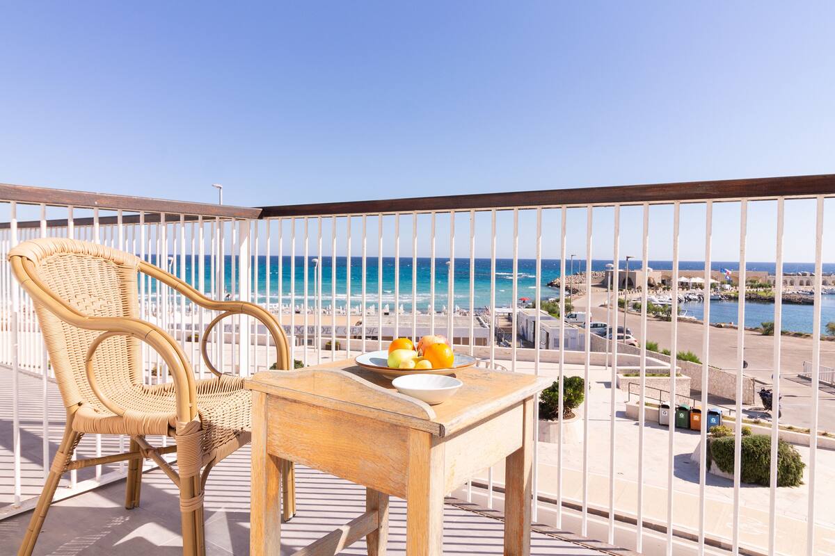 A balcony with a wicker chair and a table with fruit, overlooking a marina and the sea.