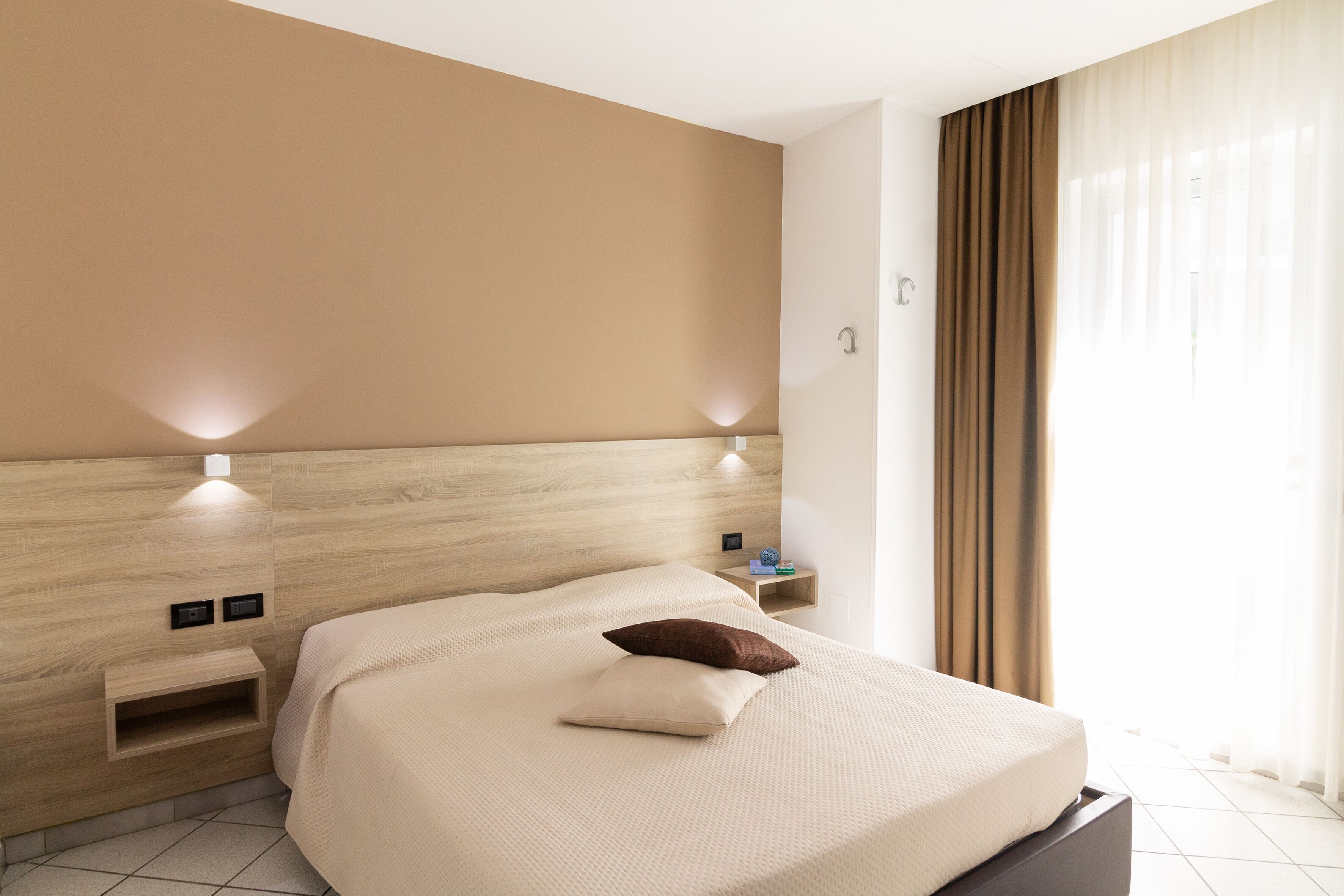 A modern hotel room with a wooden headboard, a bed with a beige cover, a nightstand, and a wall-mounted light fixture.